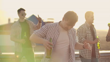 The-young-American-man-is-dancing-on-the-roof-with-his-friends-who-drinks-beer.-He-smiles-and-enjoys-the-time-in-shorts-and-a-blule-plaid-shirts-in-summer-everning.